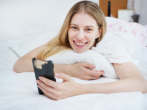 Enchanted blonde woman laughs cheerfully while talking on her mobile phone on her bed at home.
