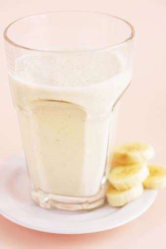 A studio DSLR close up shot of a banana and oat bran smoothie served with banana pieces on side in a big glass cup. The smoothie stands on coral coloured background.