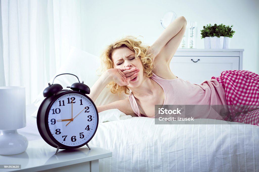 Rise and Shine Young adult woman lying on the bed in a bedroom and yawning. In the foregroung a big alarm clock showing 9 a.m. Women Stock Photo