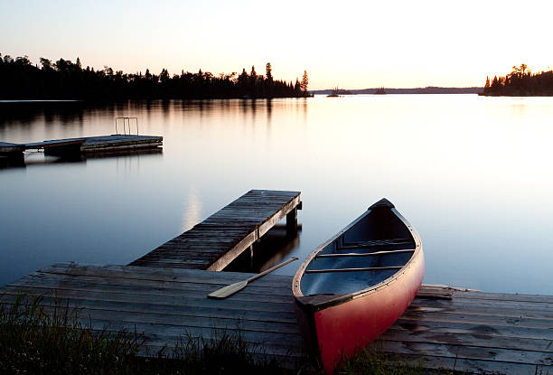 Canoe by the Dock in Muskoka Ontario Canada A classic canadiana concept. A red canoe by a dock at dusk in cottage country. Ontario, Canada. Image taken near Gravenhurst in the heart of the muskoka region. This region is popular for tourists and weekenders from Toronto. This is a peaceful scene with red canoe, rustic wooden dock, beautiful calm water, classic sunset, and oar resting on the dock. Time to go paddling!  holiday camp stock pictures, royalty-free photos & images