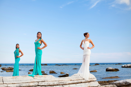 Three beautiful ladies in the evening dresses standing on the rocks on the sea lagoon