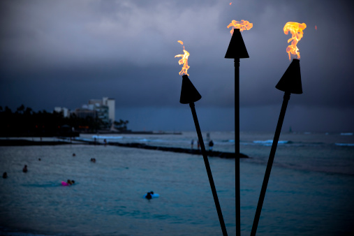 Late evening over Waikiki Beach with overcast sky. Focus on three flaming torches in a foreground. There are late swimmers in a distance.