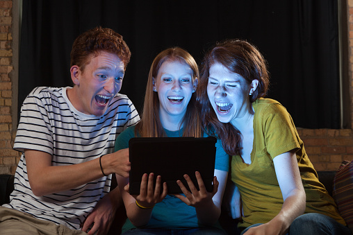 Three young college students, young adult friends watching a video streaming a funny comedy movie on a tablet computer, laughing together at humor and jokes.