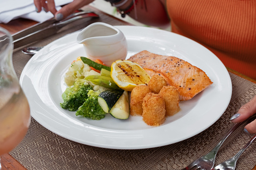 Close-up shot of unrecognizable woman eating salmon steak with vegetables and potato croquette. Her hands cutting the salmon with table knife.