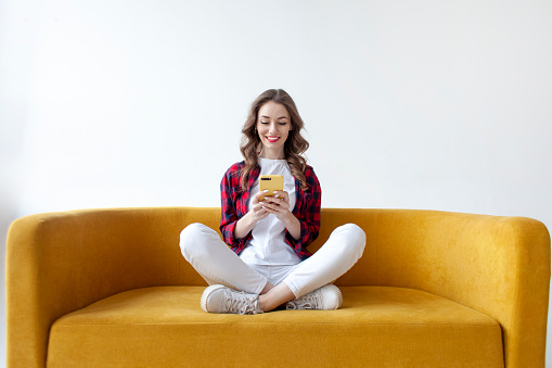 young cute girl use smartphone on soft comfortable sofa, woman is typing message on mobile phone online on yellow couch on white isolated background