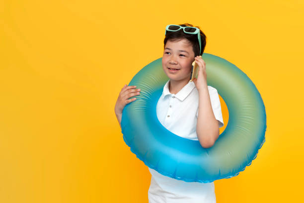 黄色の隔離された背景に電話で話しているインフレータブル水泳リングを持つ10歳のアジアの小さな男の子 - 10 11 years little boys glasses portrait ストックフォトと画像