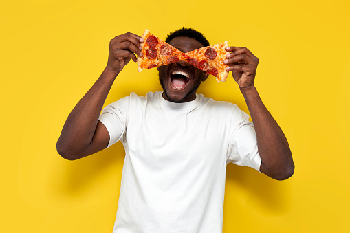 african american man in white t-shirt holds two pieces of pizza in front of his eyes and screams with his mouth open on yellow isolated background, funny guy eats fast food and shows glasses from food