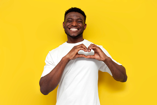 joyful african american man in white t-shirt shows heart with his hands on yellow isolated background, young guy shows gesture of love and romance and smiles