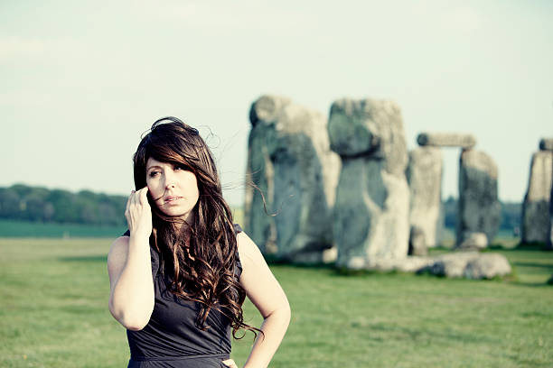 Woman in dress and long brown hair stands in front of Stonehenge at dusk. Wind blows her hair as she looks to the side. Warm colors of dusk.