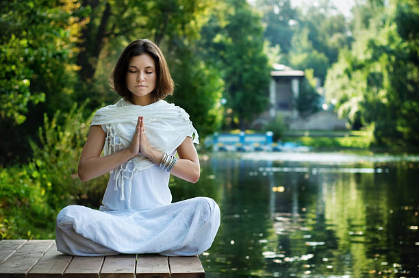 Young woman practicing yoga "Young woman sitting in lotus position near the water, practicing yoga and meditating." chanting stock pictures, royalty-free photos & images