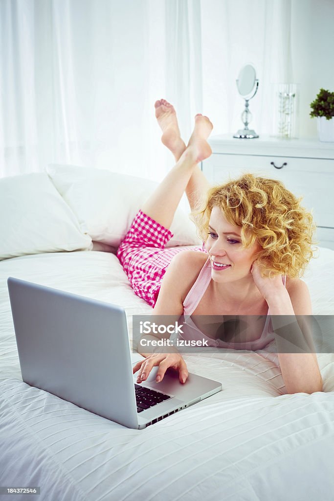 Using computer in a bed Smiling young adult woman lying on stomach on a bed and using laptop, looking at the screen. 20-24 Years Stock Photo