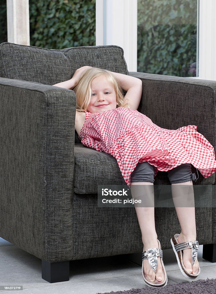 lazy girl in armchair grinning lazy blond girl in red sleeveless dress lying in armchair in livingroom looking at the camera Girls Stock Photo