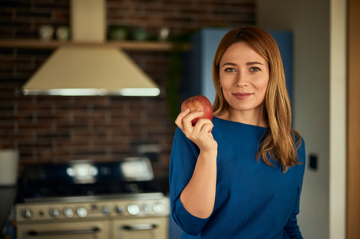 A blonde woman is in the kitchen, holding an apple, and smiling for the camera.
