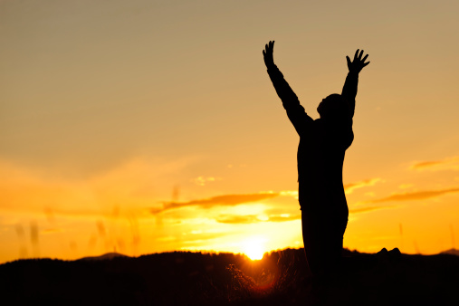 free woman at sunset praying; arms raised.