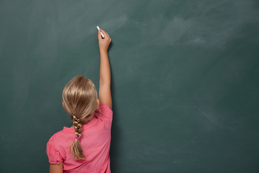 Back view of little girl with blonde hair writing on blank blackboard.She is wearing a pink dress and stands on the left side of frame.The black board is blank.Studio shot with a full frame DSLR camera.
