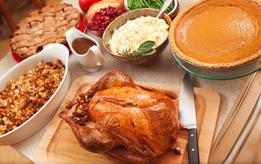 Thanksgiving or Christmas holiday dinner foods featuring roast turkey, cranberry sauce, stuffing, gravy, mashed potatoes, squash, green beans, and sweet potatoes vegetable side dishes, with pumpkin and apple pie desserts. The poultry rests on a kitchen counter cutting board, prepared for a party. Abundant fall meals are USA celebration traditions.