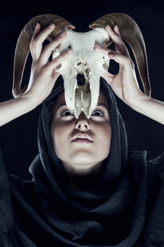 woman in the hood raises sheep's skull over his head. Black background. Looking up.