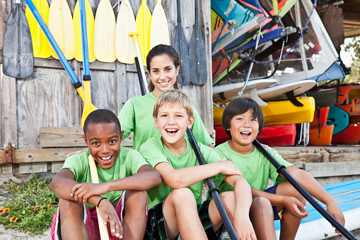 Teenage girl (17 years) with boys (8-9 years) sitting on paddle board at water sports equipment shack.