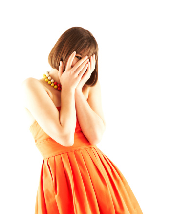 Portrait of a young woman peeking through covered face wearing orange dress over white background