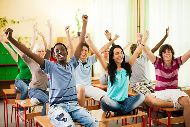 Large group of students sitting on desks with raised arms. Large group of successful teenage students is sitting on their desks in the classroom with raised arms. teenage high school girl raising hand during class stock pictures, royalty-free photos & images