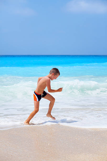 Young little boy on the beach Little boy enjoying the beautiful beach, smiling and running in front of large wave. Photo was taken on one of the most beautiful beaches in the world. The Egremni beach. Lefkada island in Greece. egremni beach lefkada island greece stock pictures, royalty-free photos & images