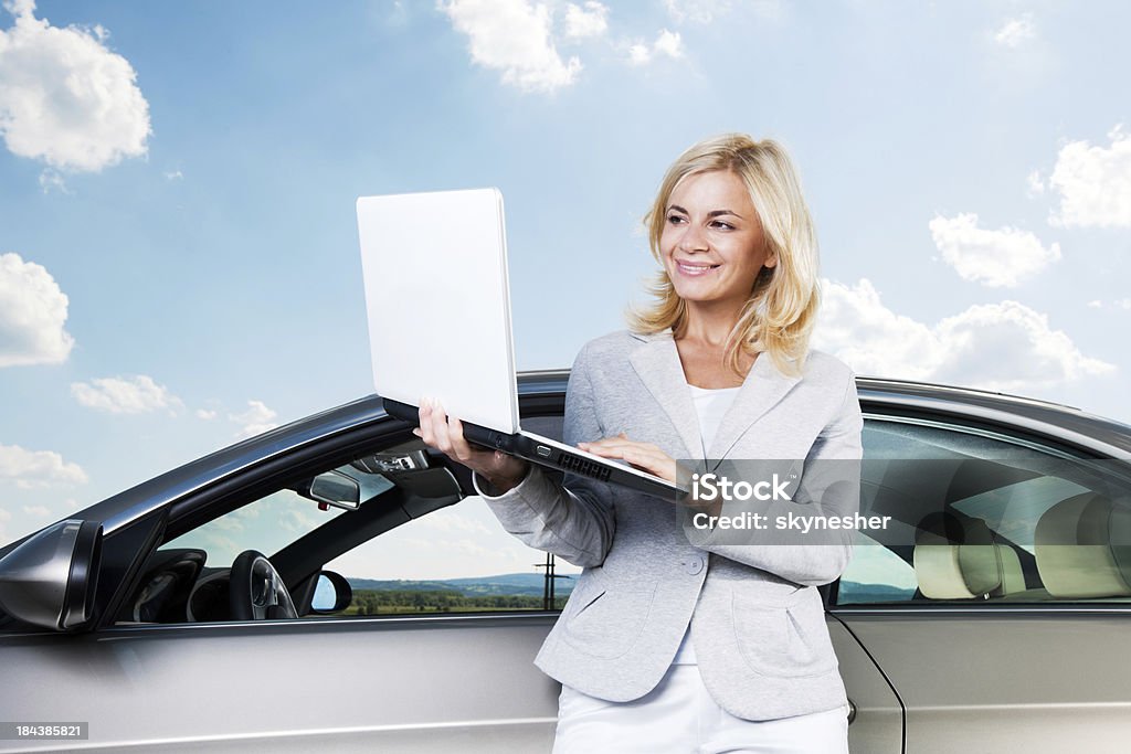 Donna sorridente utilizzando il proprio computer portatile contro il cielo e cloud. - Foto stock royalty-free di Automobile