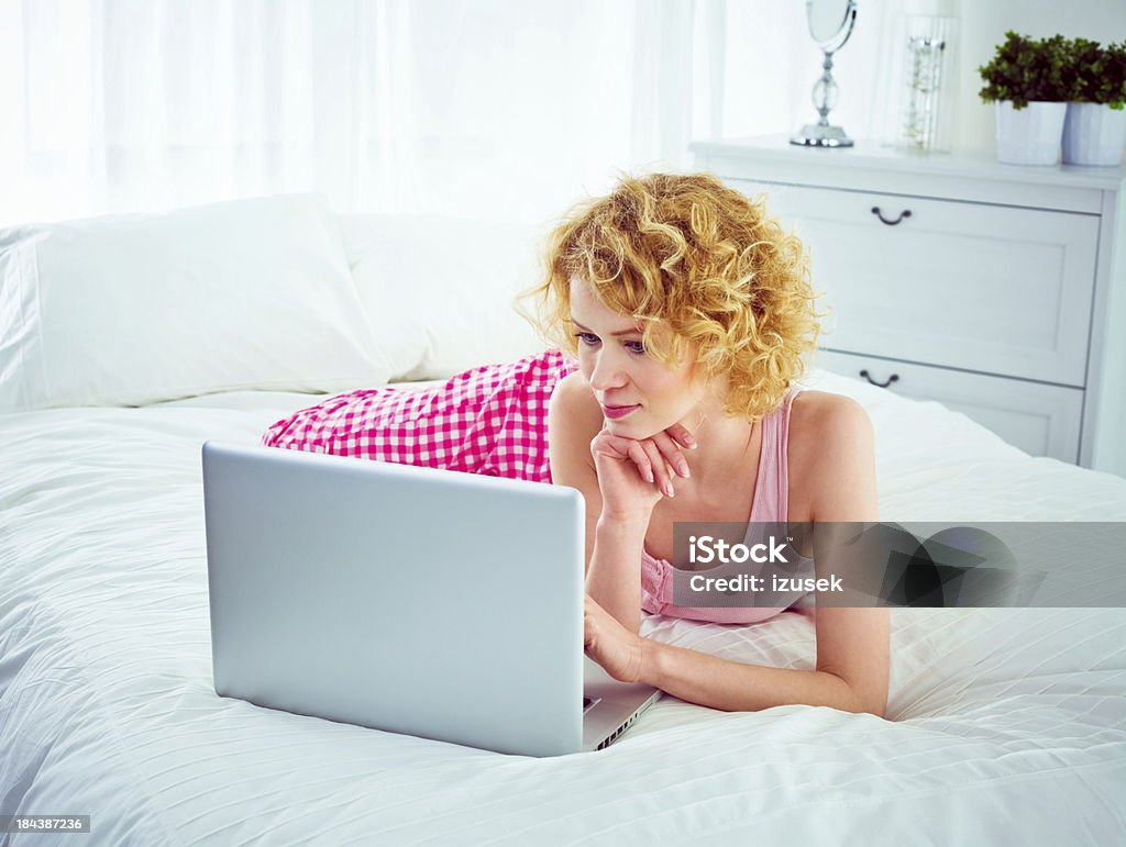 Using computer in a bed Smiling young adult woman lying on stomach on a bed and using laptop, looking at the screen. 20-24 Years Stock Photo