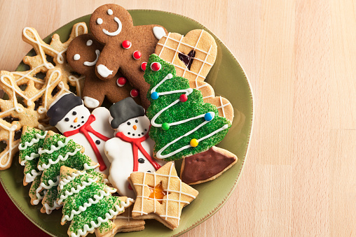 A plate filled with Christmas cookies decorated with frosting and icing for winter holiday celebrations. The baked dessert collection of sweet food features Christmas tree, gingerbread man, snowman, star, heart, and snowflake shapes against the green background of a ceramic plate. The light wooden table area allows for copy space.