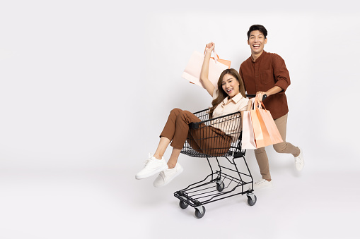 Happy Asian couple holding shopping bag and pushing shopping trolley isolated on white background