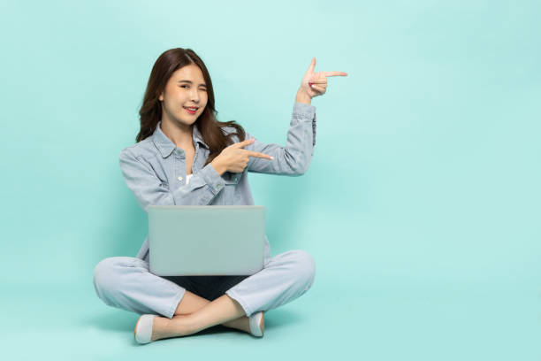 istock Happy young Asian woman freelancer pointing and using laptop computer and sitting on floor isolated on green background 1843900610