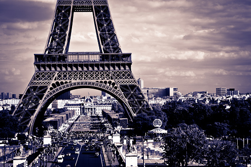 DSLR picture of the Eiffel tower in Paris on a cloudy day of summer, taken from the Trocadero. Cars and tourists are visible around the tower. In the background there are other office buildings. A purple effect was applied to the picture.