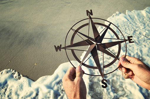 Hands hold rusty old compass over smooth sand with waves rushing ashore on beach