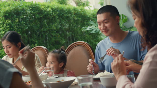 Asian family eating chinese food and having fun sitting at dining table at backyard outside home. Multi-generation family enjoying spending together.