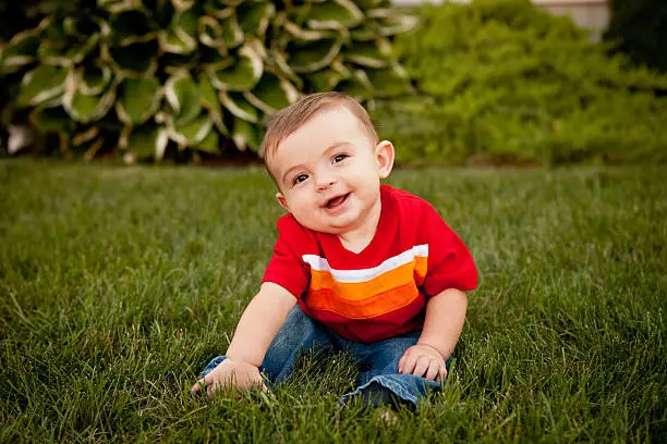 Color photo of a happy six-month-old baby boy sitting up and smiling in an outdoor setting.