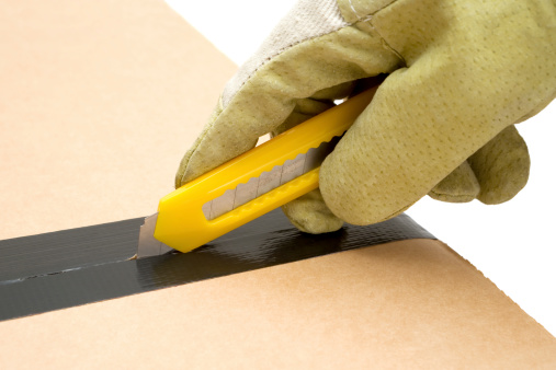 Person in work gloves using a box cutter knife to open a sealed cardboard box. White background.