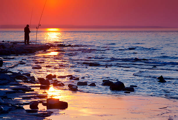 Sunset and Fisherman "The sunset lit sky ignites the rocky shore of Georgian Bay and silhouettes a fisherman at Craigleith Provincial Park near Collingwood, Ontario.  The shale that forms the shore in this part of  Georgian Bay is studded with invertebrate fossils believed to be more than 400 million years old." sea fishing stock pictures, royalty-free photos & images