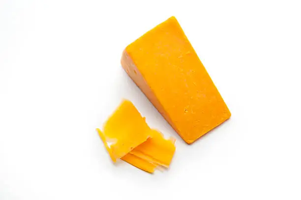 A block and some shaved slices of cheddar cheese on a white studio background.  