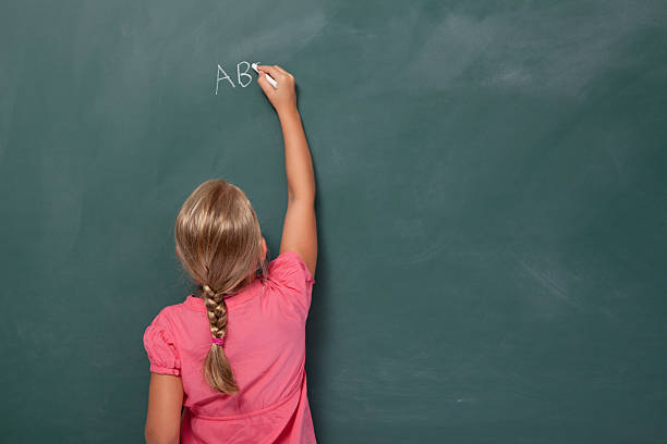 Back view of little girl with blonde hair writing alphabet on blank blackboard.She is wearing a pink dress and stands on the left side of frame.The black board is blank.Studio shot with a full frame DSLR camera.