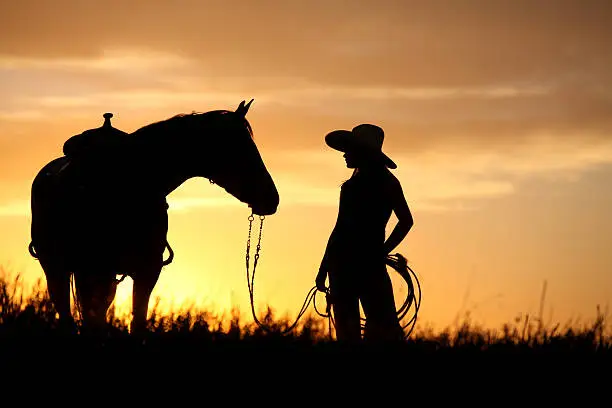 cowgirl looking at her horse with the sun setting in the background.