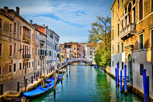DSLR picture of a canal in the city of Venice, Italy.  The walls of the buildings are multi coloured in bright colours.  It's a nice summer day, the sky is cloudy but the blue sky is visible. There is reflections of the buildings into the canal and blues gondolas are in the foreground.