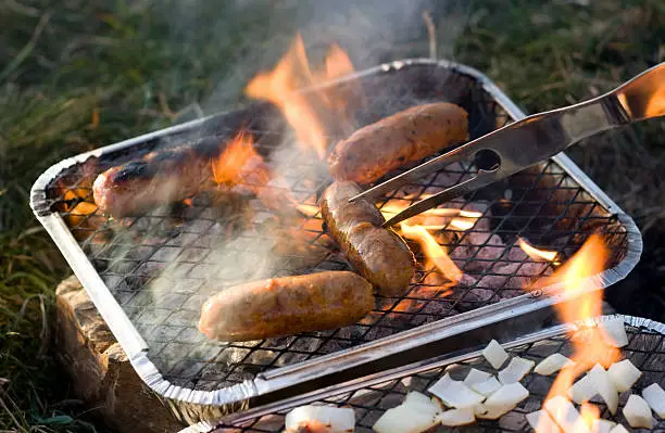 "Sausages cooking on a disposable barbecue outdoors. Main focus on sausage being turned, soft focus on background."