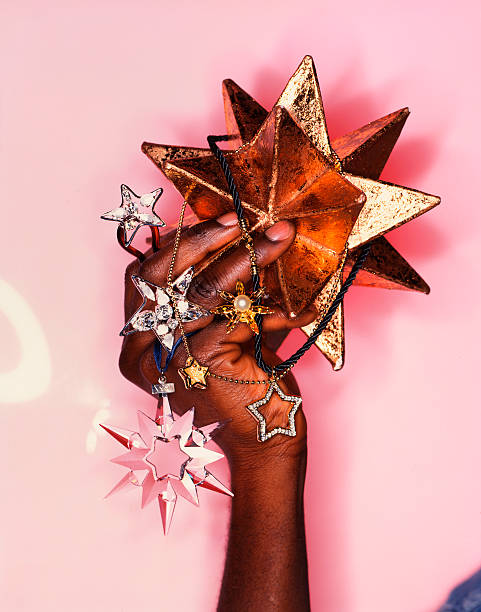 A black hand holding several Christmas stars and other decorations on a pink background shot with a ringflash