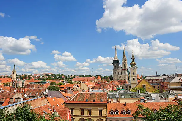 A view across numerous rooftops in Zagreb showing the cathedral Kaptol as a prominent feature to the right of the center.  The cathedral is the tallest building appearing in the skyline and shows two peaks.  A building with a clock on the tower has a pointed peak and also appears on the right side.  The sky is a bright blue color with scattered clouds.  Most of the rooftops are red and contrast with the beige and gold colors of the cathedral.  The tops of trees appear amongst and above some of the rooftops.
