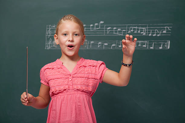 Little Girl Directing Chorus For Folk Music Before Blackboard Little cute blonde haired girl is standing in front of blackboard and directing chorus.A folk music song notes written on blackboard.The model is looking at the camera.She is wearing a pink dress and located on left side of frame.She is holding a stick in right hand.Studio shot with a full frame DSLR. conductors baton photos stock pictures, royalty-free photos & images