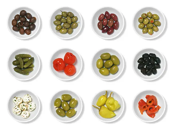 An assortment of olives and antipasto isolated on white.  From left to right, top to bottom, they are: pitted black olives, Greek olives, Kalamata olives, cocktail olives with pimentos, cornichons, sweet pickled red peppers, large green olives, dried olives, marinated mozzarella, feta stuffed olives, pepperoncinis, and roasted red peppers (pimentos).