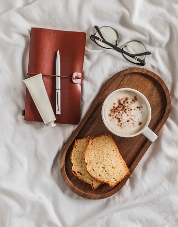 A cozy morning - a cup of coffee, pieces of cupcake on a wooden tray, a notebook, hand cream, glasses on white sheets in bed