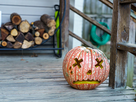 An artistically carved pumpkin with a unique pattern sits on a wooden porch, signaling the start of the festive Halloween season with logs stacked in the background.