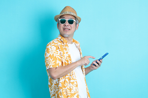 Portrait of Asian energetic senior man holding a mobile phone isolated on the blue background with travel clothes. mature man, Asian man. Elderly, Holiday, Travel. studio shot. Multiple emotions.