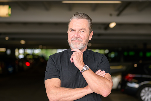 Attractive gray-haired man with a black T-shirt wears a stylish watch and looks friendly at the camera, in front of a car park