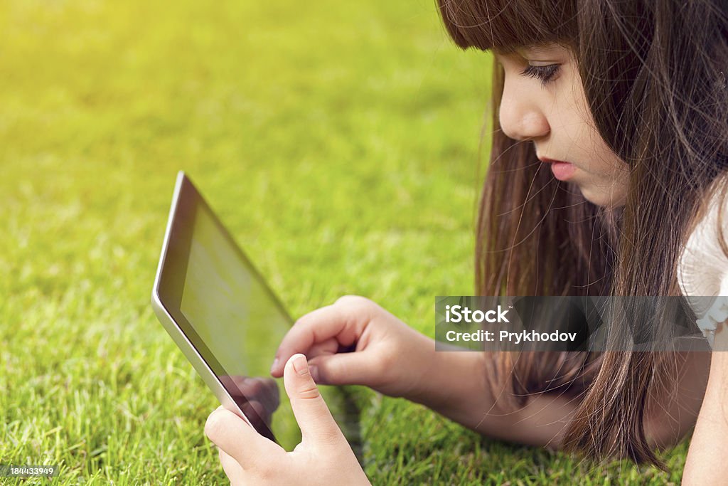 Piccola ragazza sdraiata sull'erba e tocchi lo schermo del tablet - Foto stock royalty-free di Attrezzatura